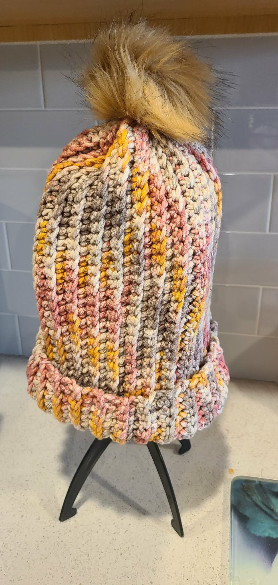 Colorful crocheted adult beanie hat with a faux fir pom-pom on a stand against a tiled wall.
