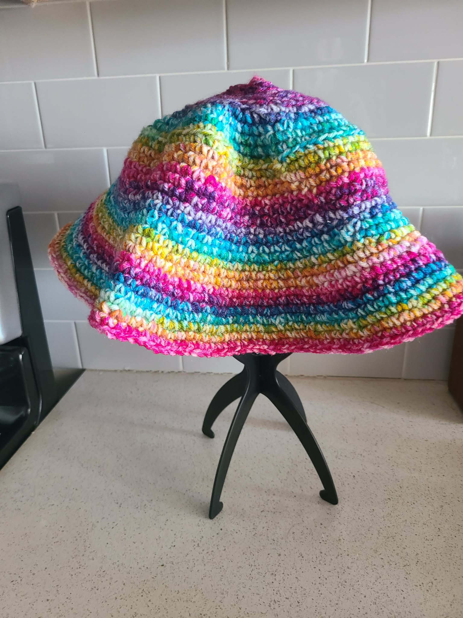 Colorful crocheted bucket hat on a small black stool against a tiled wall.